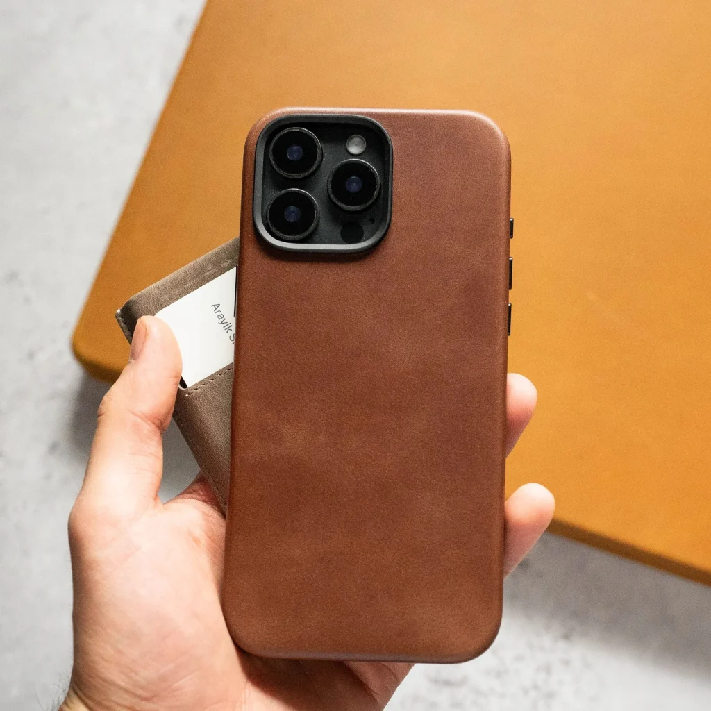 در تصویر یه دست میبینیم که قاب گوشی چرمی رو گرفته که زمینه تصویر یه بکگراند نارنجی و طوسی روی زمین هست که یه طرح فانتزی جذاب ساخته